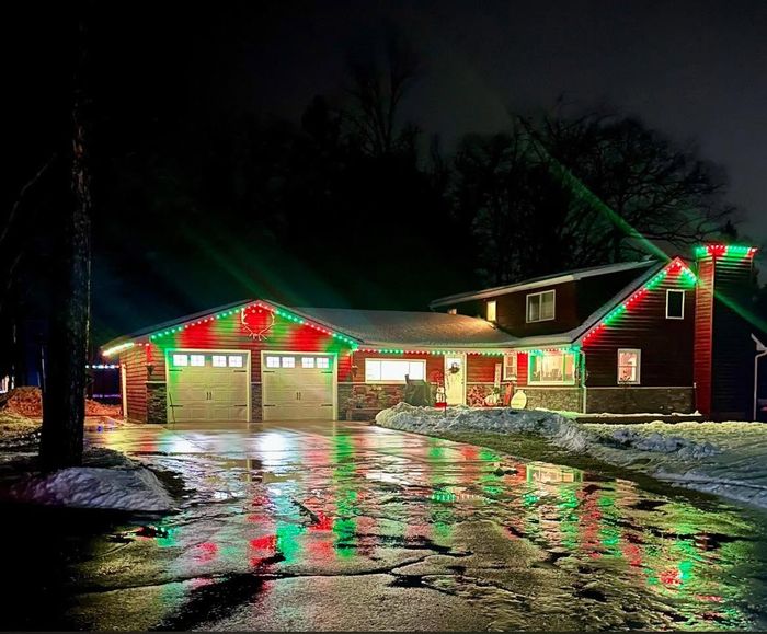 Cabin-style home with red and green Trimlight reflecting off a wet driveway at night