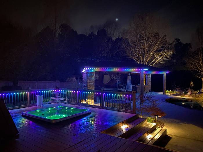 Backyard deck with hot tub and pergola lit with rainbow Trimlight along the railing